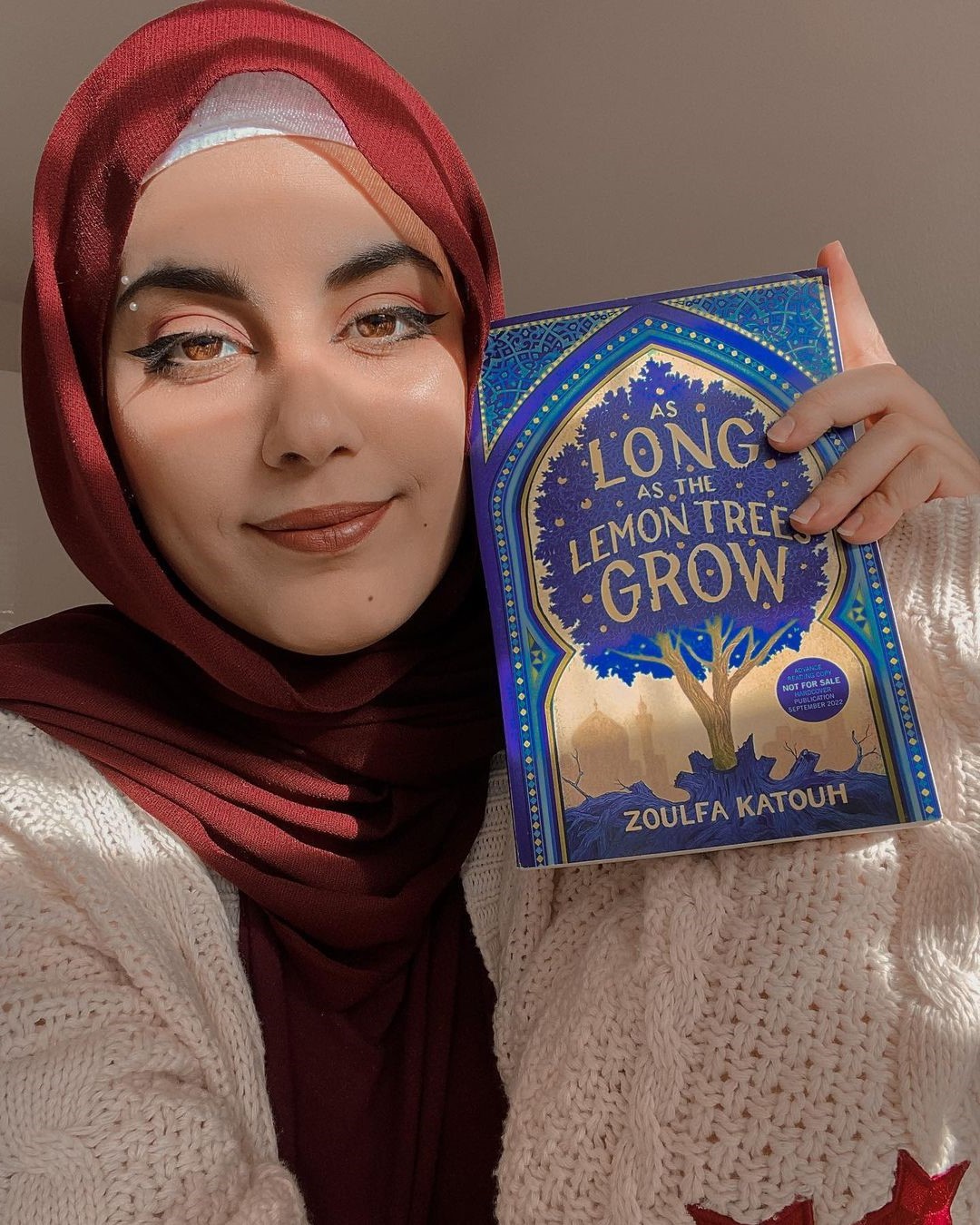Portrait of Zoulfa Katough posing with the book "As Long As the Lemon Trees Grow". She is a young woman in a red hijab with pointed eyeliner.