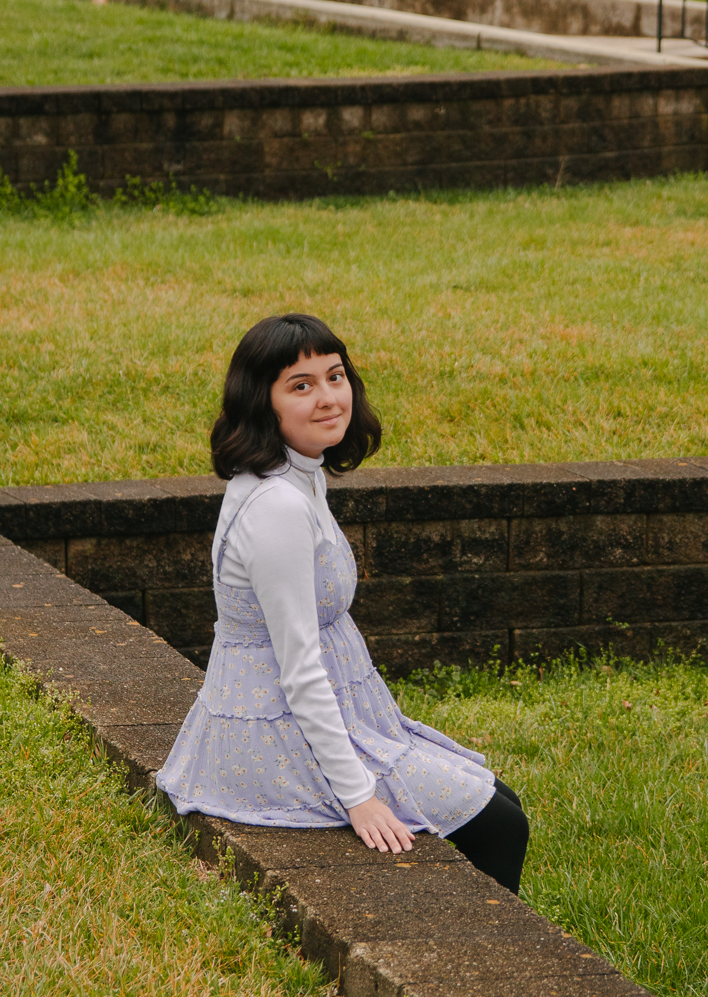 Photo of Grace sitting on a ledge in a grass field. She is smiling softly, has short black hair in a bob and is wearing a blue dress.