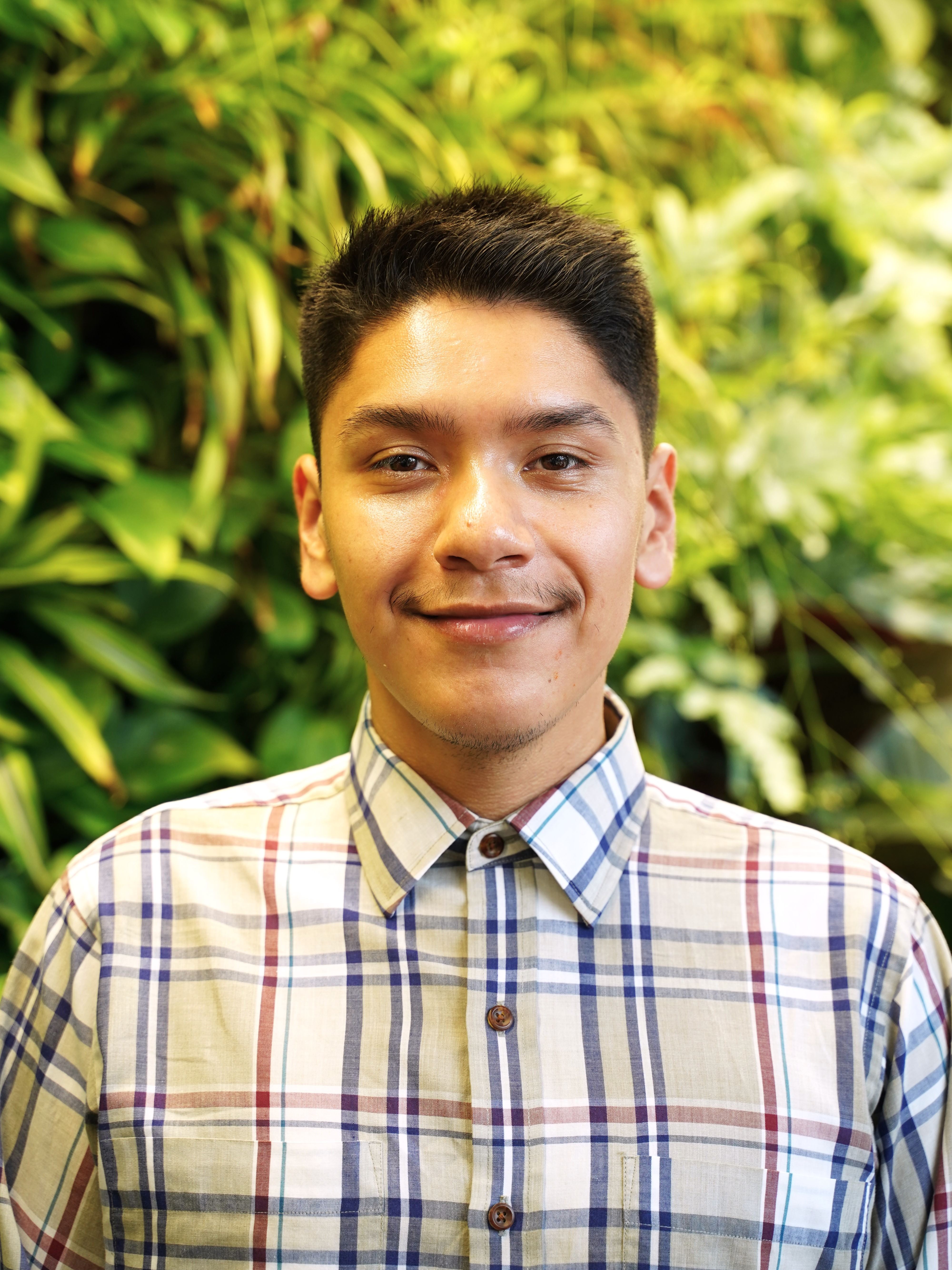 A Latino person with short Black hair stands in front of a plant wall, looking at the camera. 