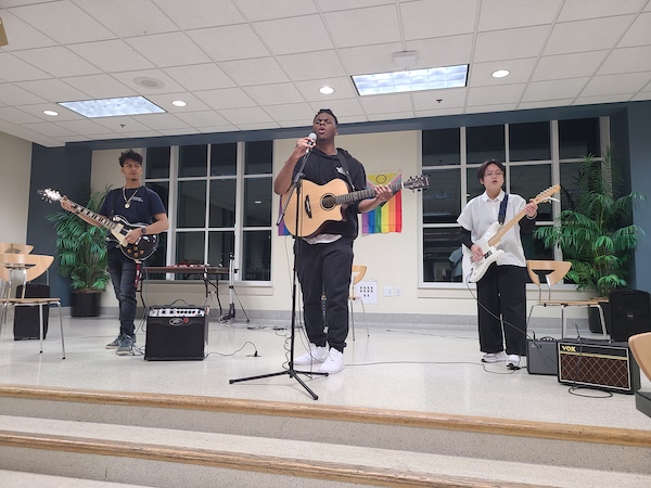 A band is performing. All three members are holding guitars. The middle figure is holding a microphone on a stand and singing into it.