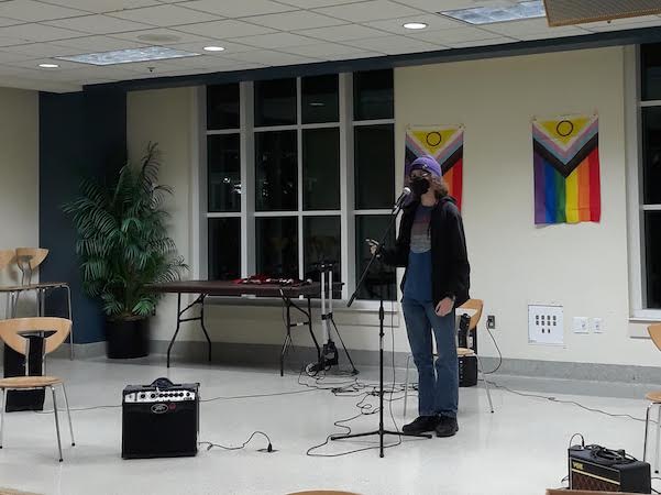 A figure is standing on a stage speaking into a microphone. Their face is obscured by their curly hair a black face mask. Two Pride Progress flags hang on the wall behind them.