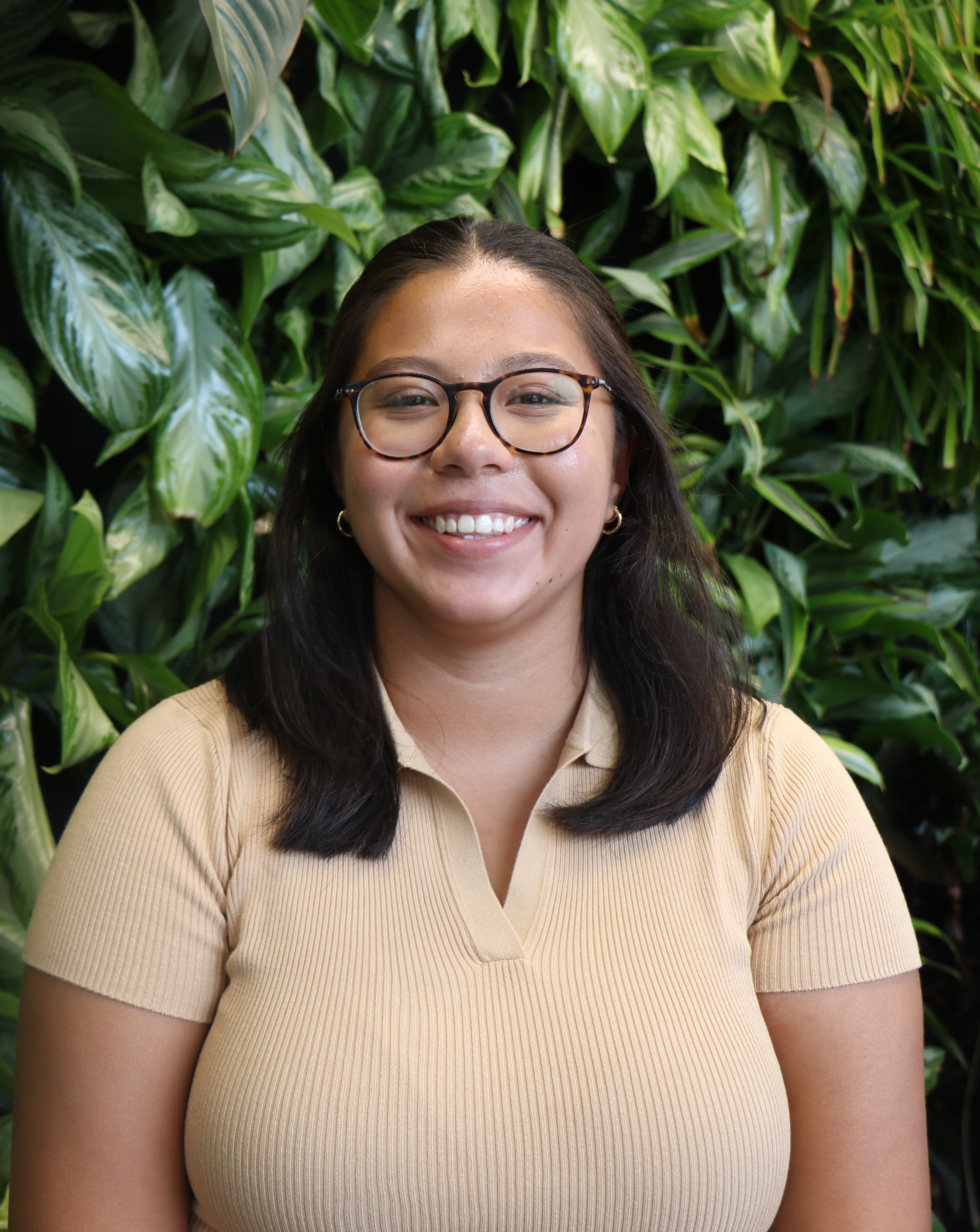 A Latina person with long dark brown hair wearing glasses, standing in from of a green wall smiling into the camera