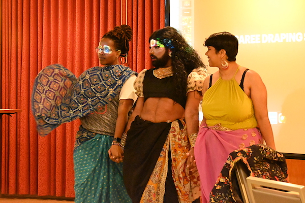 three figures wearing flowy garments draped around themselves are holding hands and posing in front of a projector screen and red curtains