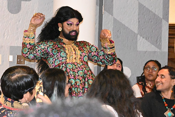 A figure in drag makeup, adorned in gold jewellery, and wearing a pink and green garment is holding up their hands while the faces and heads of people around are turned to look at them