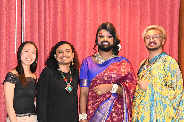 Four figures are standing next to each other and smiling: a person in a black lace-hemmed top with dark hair; a person wearing a chunky multicolored necklace over a black top with shoulder length hair; a person wearing a blue and red saree with gold and pearl jewellery and drag makeup; and a person with light hair wearing a yellow and blue saree.