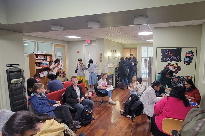 Students sitting and standing in a casual lounge/study space indoors, chatting, eating, and using phones and laptops.