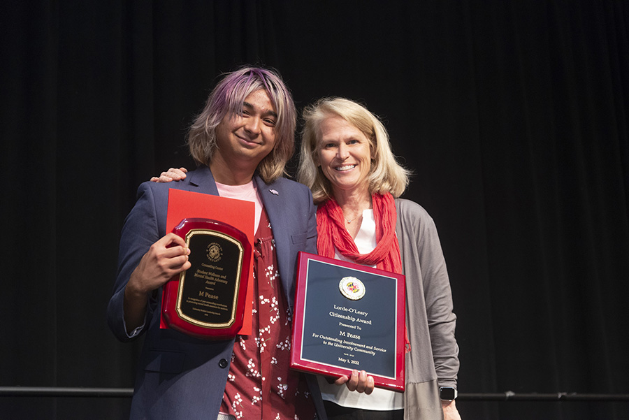 figure with purple hair smiles and holds up a wooden plaque of the award in one hand and a certificate in the other, standing next to figure with blond hair 