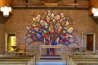 Interior of a small brick chapel sanctuary featuring a vibrant, multi-colored tree mosaic on the back wall.