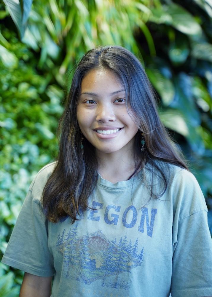 Person with long hair is smiling at the camera. They are wearing a forest green graphic tee.