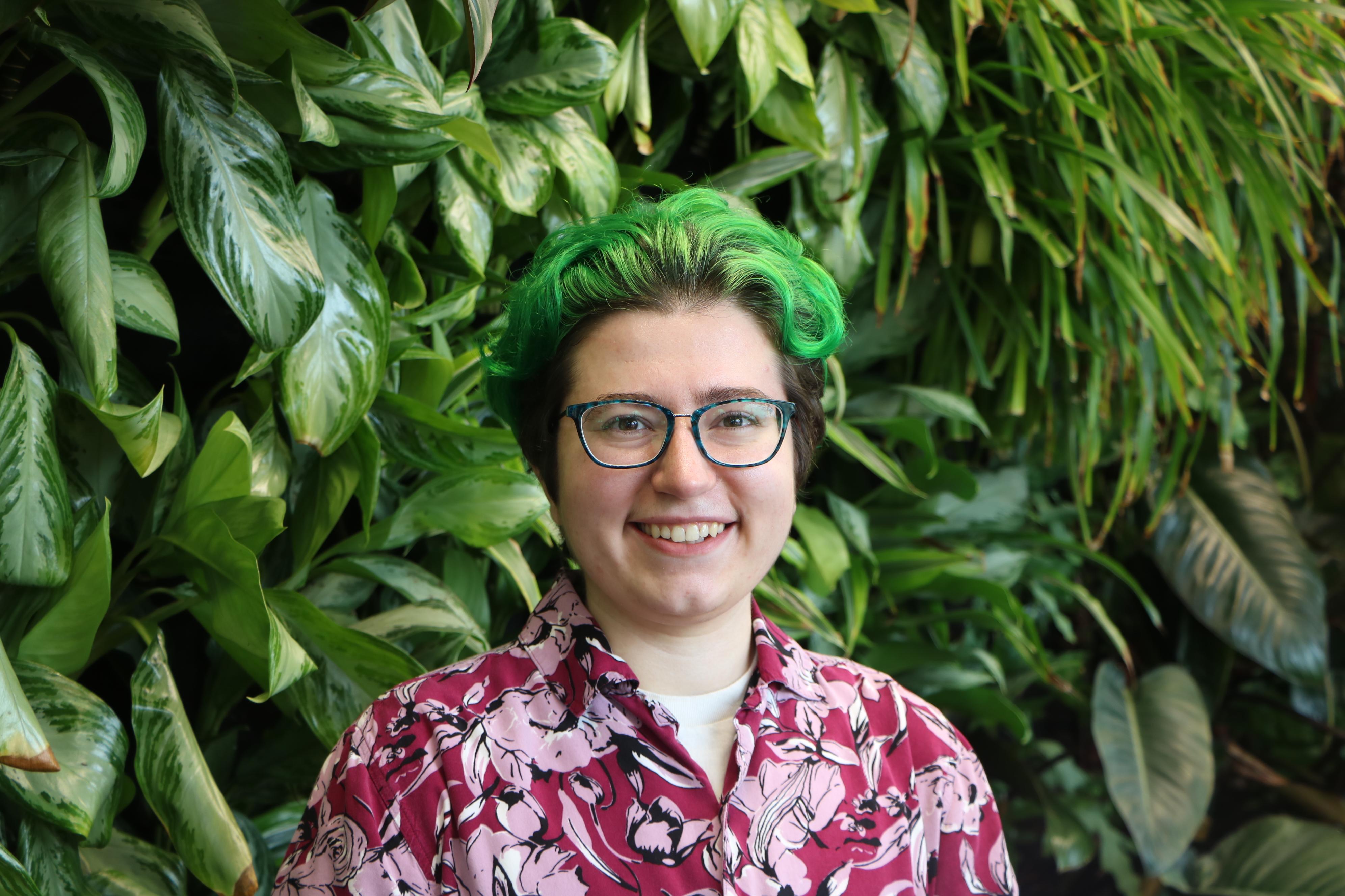 A picture of LGBTQ+ Graduate Coordinator Freddie Dufresne standing in front of the plant wall in the STAMP Student Union. They are smiling, have glasses and bright green short hair, and are wearing a magenta floral button-up.