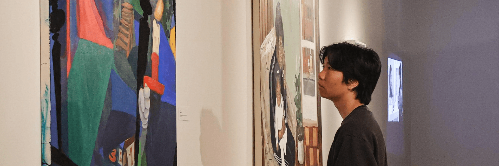Image shows a young man looking at a painting by Lolo Gem. The painting depicts a cartoon-ish stair labyrinth in dark shades of blue and green. A disembodied gloved hand holds a lit candle as if going up the stairs.