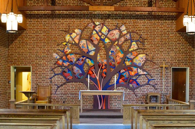 Interior of a small brick chapel sanctuary featuring a vibrant, multi-colored tree mosaic on the back wall.