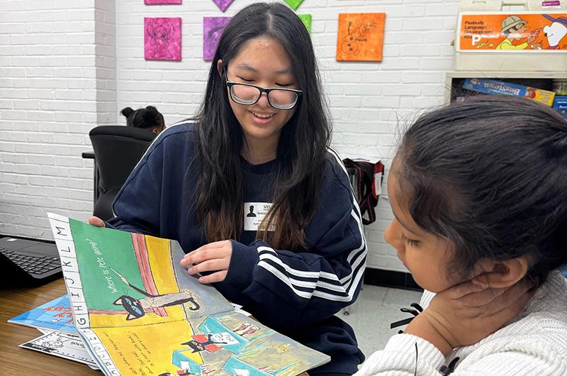 A university student smiling while reading a children's book to a young girl in a classroom setting.