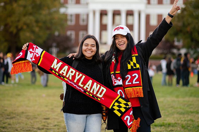 Two smiling students on McKeldin Mall holding up a red University of Maryland spirit scarf during Terp Carnival.