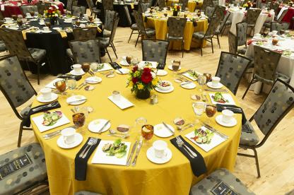 A large ballroom set up with multiple round dining tables, featuring yellow, black and red linens and floral centerpieces.