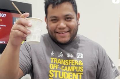 A smiling student holds up a small paper cup while wearing a grey "Transfer & Off-Campus Student Life" t-shirt.