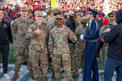 Military service members in camouflage uniforms give a thumbs-up on a football field during a stadium event.